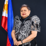 Philippine Foreign Secretary Ma. Theresa Lazaro standing beside the Philippine flag in a black and white embroidered formal outfit.