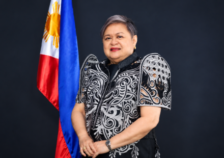 Philippine Foreign Secretary Ma. Theresa Lazaro standing beside the Philippine flag in a black and white embroidered formal outfit.