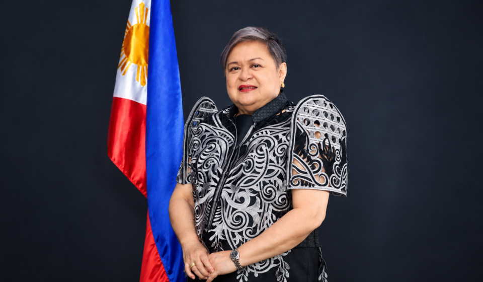 Philippines Summons Chinese Ambassador Over Remarks Seen as Threat of Force Philippine Foreign Secretary Ma. Theresa Lazaro standing beside the Philippine flag in a black and white embroidered formal outfit.