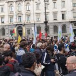 A crowd of protesters holding flags and banners in a public square in Milan, Italy, in front of a historic building.