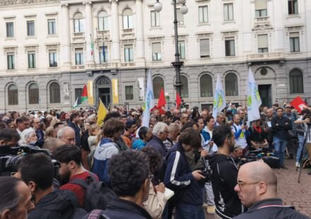 A crowd of protesters holding flags and banners in a public square in Milan, Italy, in front of a historic building.