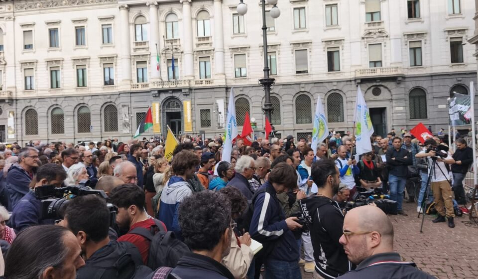 Europe Braces for Fresh Demonstrations as Milan Reinforces Security With 500 More Soldiers A crowd of protesters holding flags and banners in a public square in Milan, Italy, in front of a historic building.