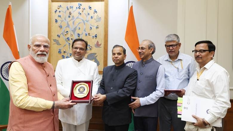 Prime Minister Narendra Modi stands with government officials holding a commemorative plaque in front of Indian national flags.
